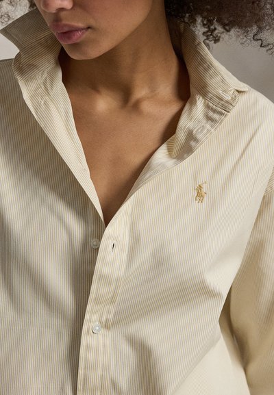 Κοντινό πλάνο ενός ατόμου που φοράει πουκάμισο με λεπτές ριγές σε απαλό κίτρινο χρώμα, με κουμπιά, και ένα μικρό κεντημένο λογότυπο στο στήθος.