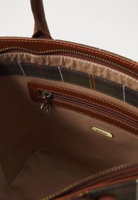 Εσωτερικό καφέ δερμάτινης τσάντας με φερμουάρ, λείο υφασμάτινο επένδυση και χρυσό λογότυπο. Πλαϊνή λεπτομέρεια καρό στην πάνω άκρη.