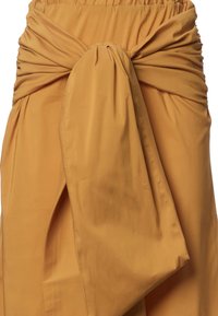 Gonna color senape con una vita arricciata, caratterizzata da un ampio dettaglio annodato sul davanti e un tessuto leggero e liscio.