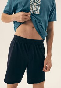 T-shirt z bawełny w kolorze teal z białym grafiką "SUNRISE SURF CLUB"; zestawione z czarnymi szortami na elastycznej talii, casualowymi i luźnymi.