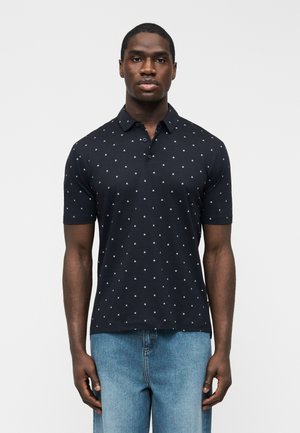 Muž v čiernej polokošeli s krátkym rukávom s bielym potlačou "AK" a svetlomodrými džínami stojí pred jednoduchým bielym pozadím.