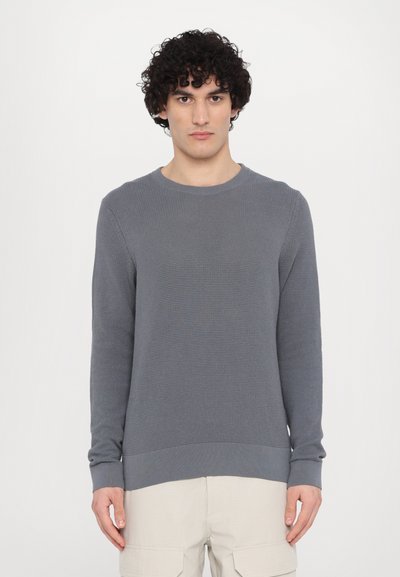 Gray textured knit sweater with long sleeves and a crew neck. Features a ribbed hem and cuffs, worn over light-colored pants.