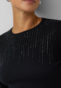 Schwarzer Strickpullover mit vertikalen Reihen aus reflektierenden Pailletten auf der Vorderseite, der ein strukturiertes Muster und einen Rundhalsausschnitt betont.