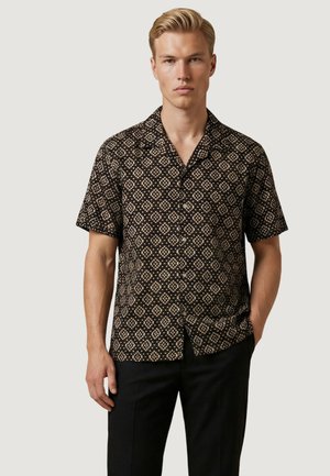 Uomo che indossa una camicia nera a maniche corte con motivo geometrico beige e pantaloni neri, in piedi davanti a uno sfondo chiaro e neutro.