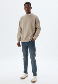 Sudadera beige de cuello redondo con mangas raglán, combinada con unos jeans azul ajustados y zapatillas blancas con suelas beige. Diseño minimalista, textura suave.