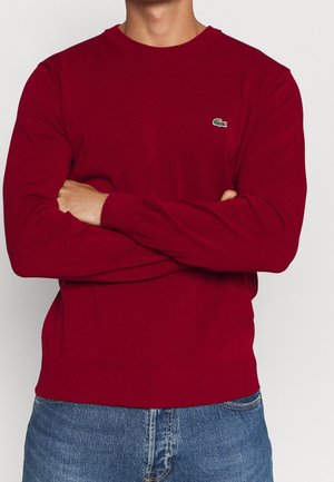 Hombre con los brazos cruzados, vistiendo un suéter rojo de manga larga liso con un pequeño logo verde de cocodrilo en el pecho y jeans azules.