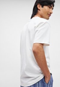 Λευκό βαμβακερό T-shirt με κοντά μανίκια, διαθέτοντας στρογγυλή λαιμόκοψη και χαλαρή εφαρμογή, προβάλλεται σε μοντέλο που φορά ριγέ μπλε σορτς.