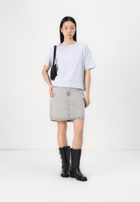 Lichtgrijs cropped t-shirt, hoge taille spijkerrok in grijs, knielange zwarte leren laarzen met gespen, en een kleine zwarte crossbody tas.
