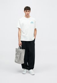 Άσπρο βαμβακερό t-shirt με τυρκουάζ λογότυπο Nike SB, μαύρο φαρδύ παντελόνι και γκρι τσάντα tote. Τα παπούτσια είναι λευκά με ορατή αερόσολα.