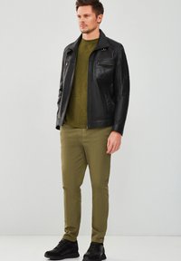 Giacca di pelle nera con tasche con zip, camicia verde oliva, pantaloni beige e sneakers neri. Design aderente, texture liscia, look moderno.