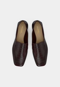 Mocassins en cuir bordeaux avec une finition texturée, design sans lacets et surpiqûres contrastées. L'intérieur présente une semelle intérieure beige avec la marque.