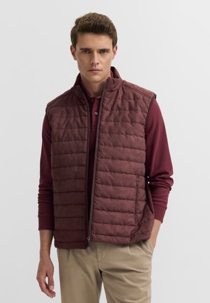 Mann trägt eine weinrote gesteppte Weste über einem weinroten Langarmshirt und beigen Hosen, steht vor einem schlichten hellen Hintergrund.