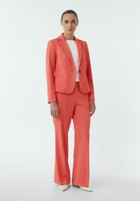Koraalblazer en bijpassende broek van zachte stof. De outfit heeft een enkele knoop en een getailleerd ontwerp, gecombineerd met een witte top en beige schoenen.