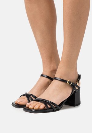 Zwarte sandalen met bandjes en een vierkante neus, gemaakt van soepel leer. Heeft een blokhak en een enkelbandje met een gouden gesp detail.