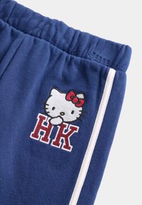Niebieskie dresowe spodnie bawełniane z elastycznym pasem, z haftowanym wzorem Hello Kitty i czerwonym napisem "HK", podkreślone białymi paskami po bokach.