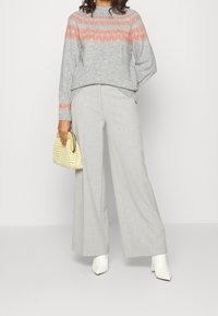 Szary sweter z dzianiny z koralowymi wzorami, w połączeniu z jasnoszarymi szerokimi spodniami i białymi botkami na szerokim obcasie. Trzyma żółtą torebkę o fakturze.