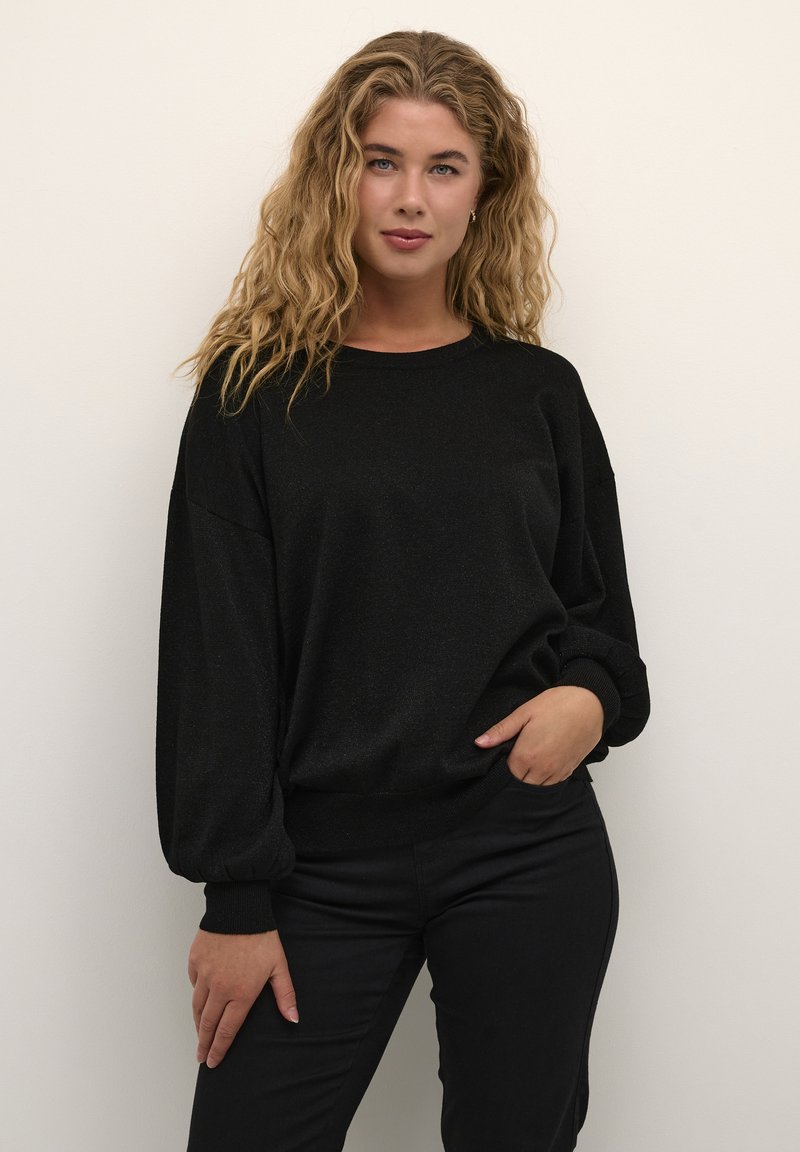 Sort sweater lavet af blødt stof, med rund halsudskæring og ballonærmer. Sættes sammen med sorte bukser, mod en neutral baggrund.