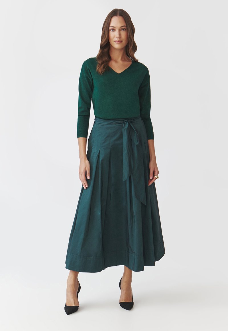 Grüner gestrickter Pullover mit V-Ausschnitt und Dreiviertelärmeln, kombiniert mit einem langen, fließenden dunkelgrünen Rock, der einen Taillengürtel und Falten hat.