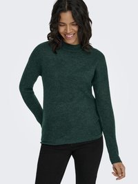 Žena sa usmieva, pozerá sa dole, oblečená v tmavozelenej pletenej mikine s dlhými rukávmi a čiernych nohaviciach, na svetlom pozadí.