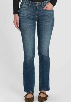Jeans in denim blu indossati da una persona con una mano in tasca, abbinati a scarpe marroni basse e una maglia nera a maniche lunghe su sfondo semplice.