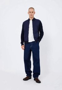 Navy Bomberjacke mit gerippten Bündchen und einem Reißverschluss, kombiniert mit dunkelblauen Jeans. Das Outfit wird mit braunen Lederstiefeln und einem weißen T-Shirt abgerundet.