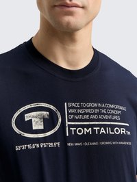 Camiseta de algodón azul marino con cuello redondo, que presenta texto impreso en blanco y un logo circular. El texto incluye coordenadas geográficas y el nombre de la marca.