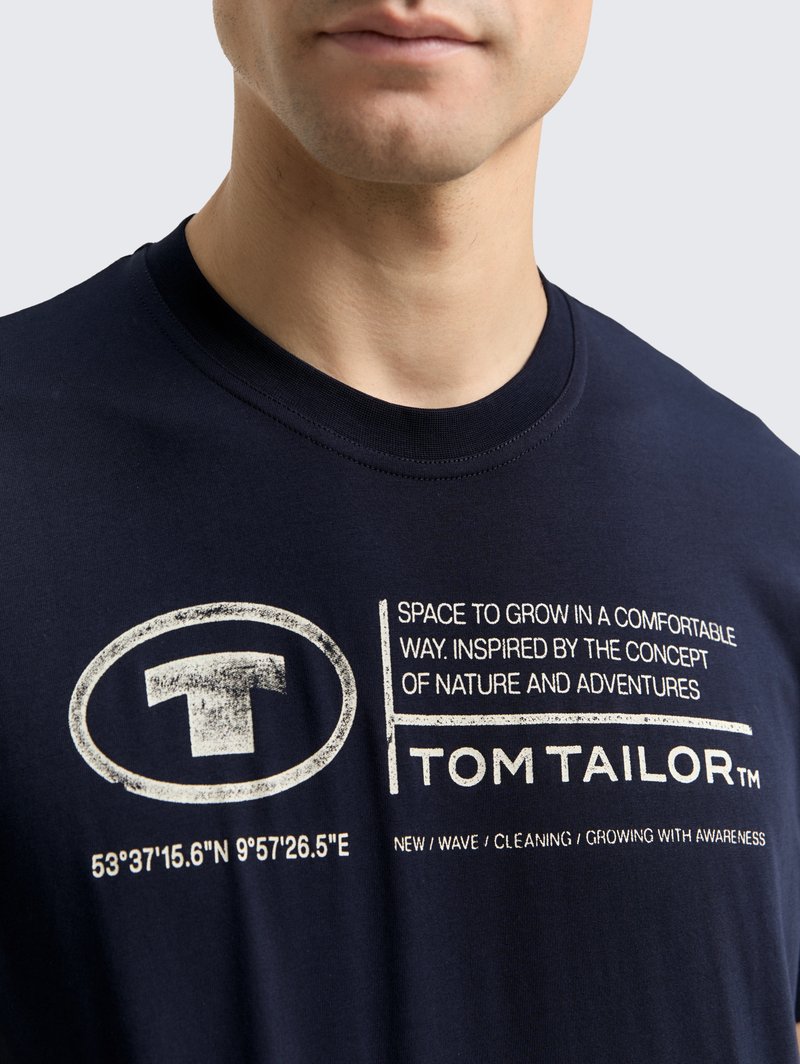 Granatowa bawełniana koszulka z okrągłym dekoltem, z białym nadrukowanym tekstem i okrągłym logo. Tekst zawiera współrzędne geograficzne oraz nazwę marki.