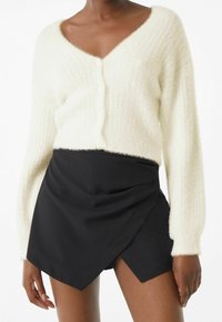 Luddig vit kofta med V-ringning och knäppning fram, tillsammans med en svart omslagstyp kjol med sned kant och mjuk textur.