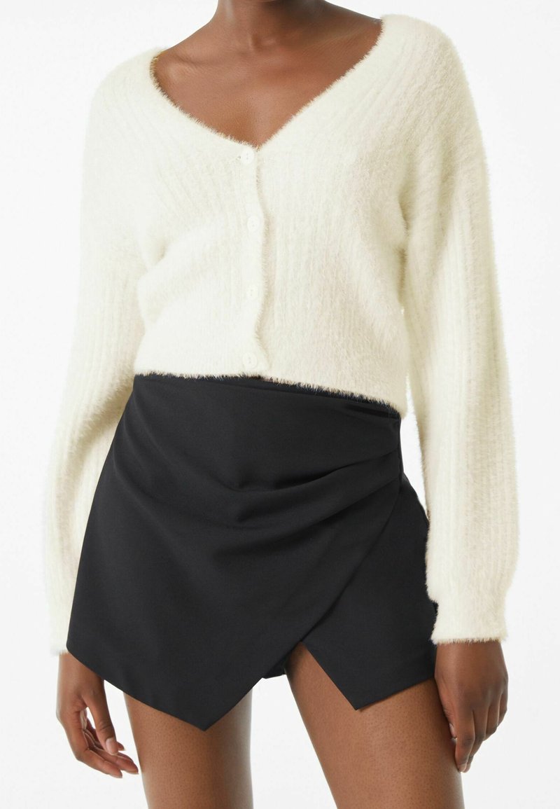 Luddig vit kofta med V-ringning och knäppning fram, tillsammans med en svart omslagstyp kjol med sned kant och mjuk textur.