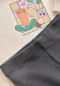 Cream-colored sweatshirt with a colorful cartoon print featuring flowers and a smiling character, paired with dark gray ribbed pants.