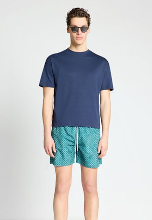 Junger Mann mit dunkler Sonnenbrille, einem einfarbigen marineblauen T-Shirt und türkisfarben gemusterten Shorts mit weißem Kordelzug, steht vor einem einfarbigen Hintergrund.
