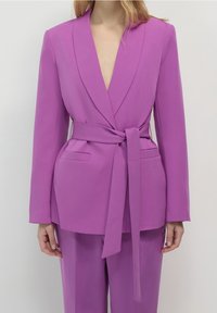 Fioletowa marynarka z głębokim dekoltem w serek, długimi rękawami, kieszeniami z przodu i paskiem w talii. Wykonana z gładkiego materiału o dopasowanym kroju.