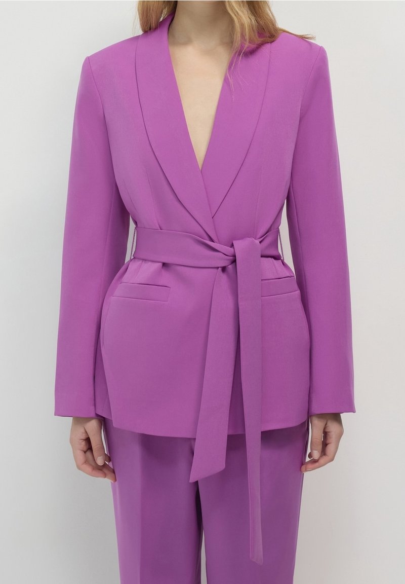 Fioletowa marynarka z głębokim dekoltem w serek, długimi rękawami, kieszeniami z przodu i paskiem w talii. Wykonana z gładkiego materiału o dopasowanym kroju.