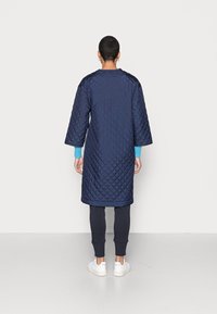 Cappotto lungo trapuntato blu navy con una vestibilità ampia e maniche a tre quarti. Presenta polsini in maglia blu e un collo rotondo. Tessuto liscio e orlo.