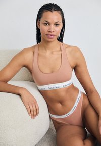 Calvin Klein bézs színű fehérnemű szett, sportos bralette-kel, logóval ellátott pánttal és passzoló magas derekú tanga bugyival. Puha, rugalmas anyag.