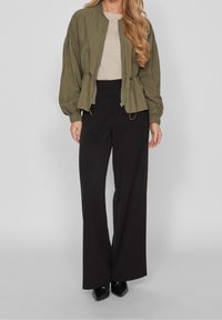 Olive zielona kurtka bombowiec z elastycznymi mankietami, zestawiona z czarnymi spodniami o szerokich nogawkach i wysokim stanie oraz czarnymi szpiczastymi butami.