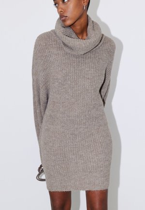 Femme portant une robe-pull beige en maille, ample, à manches longues avec un grand col bénitier, debout devant un fond blanc uni.