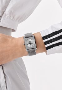 Riešas su stačiakampiu sidabriniu laikrodžiu, turinčiu pilkos audinės dirželį ir minimalistiniu ciferblatu, rodančiu valandas, minutes ir sekundes.