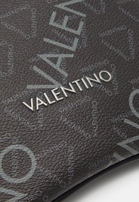 Närbild av texturerad svart läderyta med upprepande Valentino-logotypmönster och upphöjd silverfärgad VALENTINO-text.