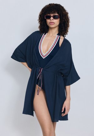 Femme portant un maillot de bain bleu marine avec un bord rouge et blanc, un paréo assorti bleu marine et des lunettes de soleil rectangulaires foncées, posant devant un fond gris.