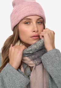 Una donna che indossa un berretto di maglia rosa e un cappotto grigio sistema una sciarpa spessa a fantasia intorno al collo su uno sfondo bianco.
