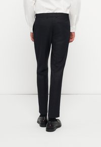 Pantalones de sastre oscuros en color azul marino con un tejido texturizado, frente plano, presillas para cinturón y un bolsillo trasero. Combinados con zapatos negros.