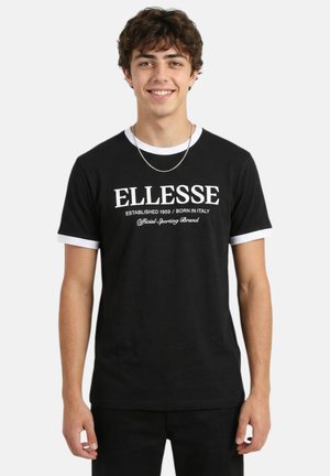 Jeune homme souriant portant un T-shirt noir avec un bord blanc et le texte « ELLESSE Established 1959 / Born in Italy » à l'avant.