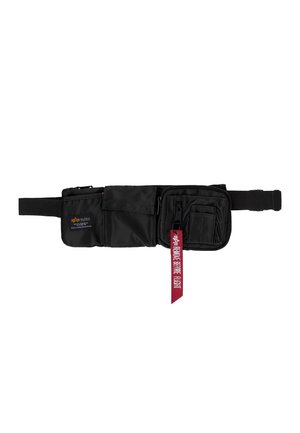 Schwarze Bauchtasche mit mehreren Reißverschlusstaschen und einem roten Stoffetikett mit der Aufschrift "Remove Before Flight", das an einem Reißverschluss befestigt ist.