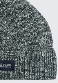 Gebreide muts in donkergroen, grijs en wit, met een textuurpatroon. Inclusief een marineblauw merklabel met witte tekst dicht bij de zoom.