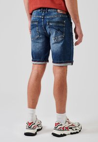 Shorts de mezclilla en un lavado azul desgastado con puños enrollados, con dos bolsillos traseros y detalles de costura. Llevados con calcetines blancos y zapatos deportivos.