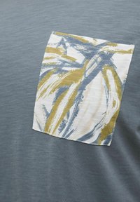 T-shirt en coton gris présentant un patch carré avec des motifs abstraits bleus, jaunes et crème. Texture lisse, manches courtes, col rond.