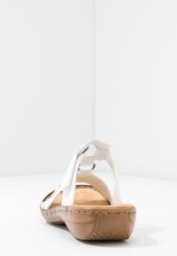 Sandale blanche avec bride arrière ajustable, semelle intérieure rembourrée et semelle en caoutchouc marron. Présente des bords cousus et une finition lisse.