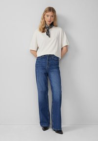 Witte oversized t-shirt, hoge taille blauwe jeans met knoop aan de voorkant, zwarte sjaal om de nek geknoopt, zwarte spitse schoenen, natuurlijk haar.