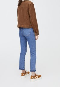 Persona in piedi, che indossa una giacca di velluto a coste marrone con colletto bianco, jeans corti blu con orli sfrangiati e sneakers a piattaforma color senape con lacci bianchi.